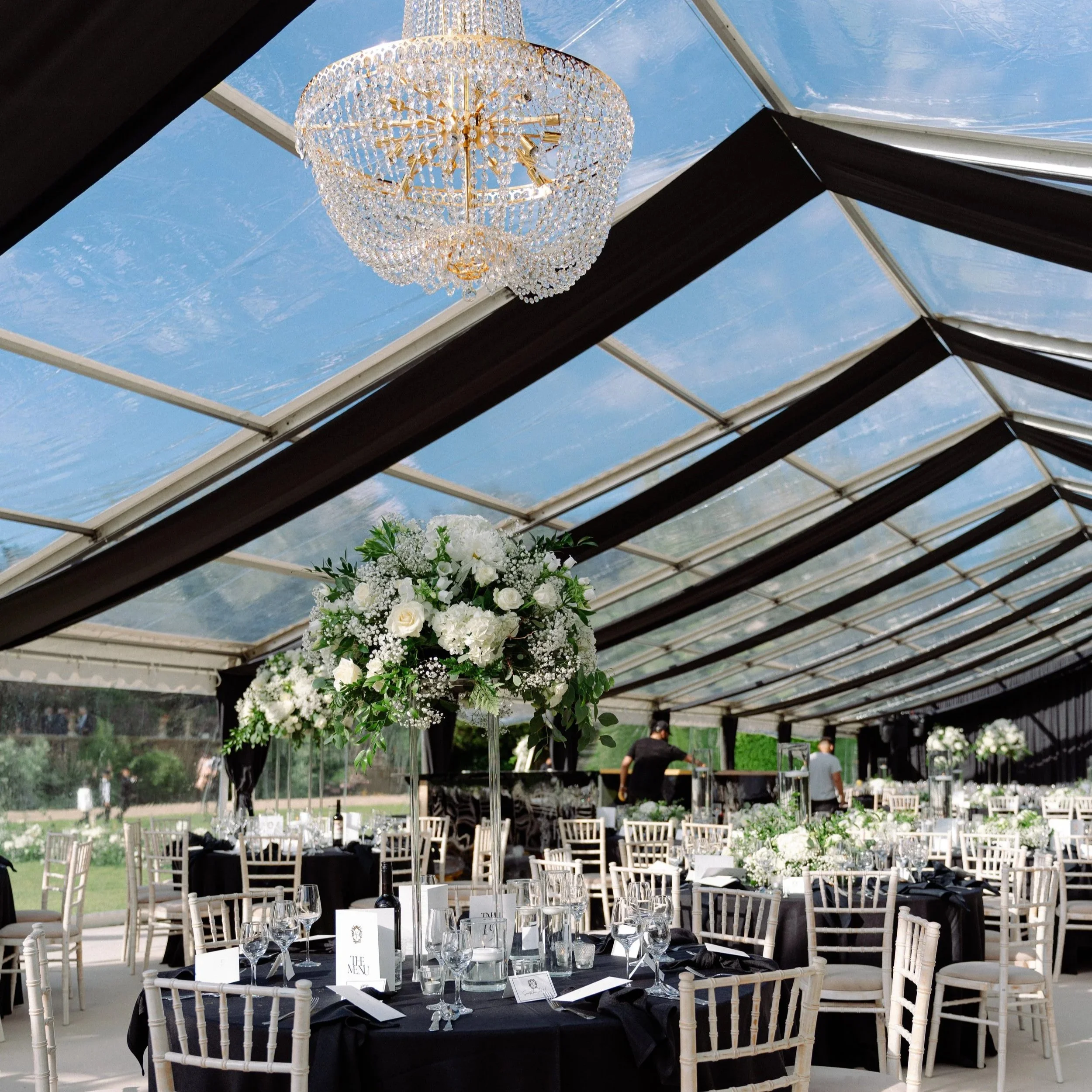 Sophisticated wedding marquee with a clear roof, black draping, and a grand crystal chandelier. Round tables are styled with black linens, elegant glassware, and tall floral centerpieces of white roses and greenery. Neutral Chiavari chairs complete t