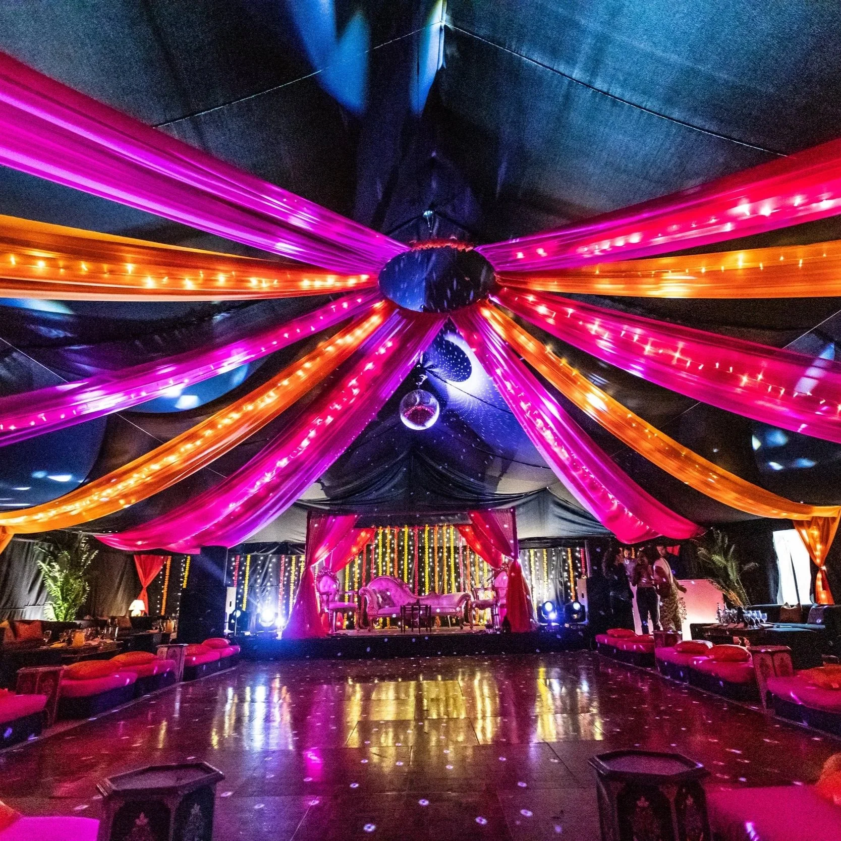Vibrant Indian wedding marquee draped with rich pink, orange, and purple fabric illuminated by fairy lights. The dramatic tent interior features a central dance floor, low seating with bright cushions, and a stage framed with red drapes and decorativ
