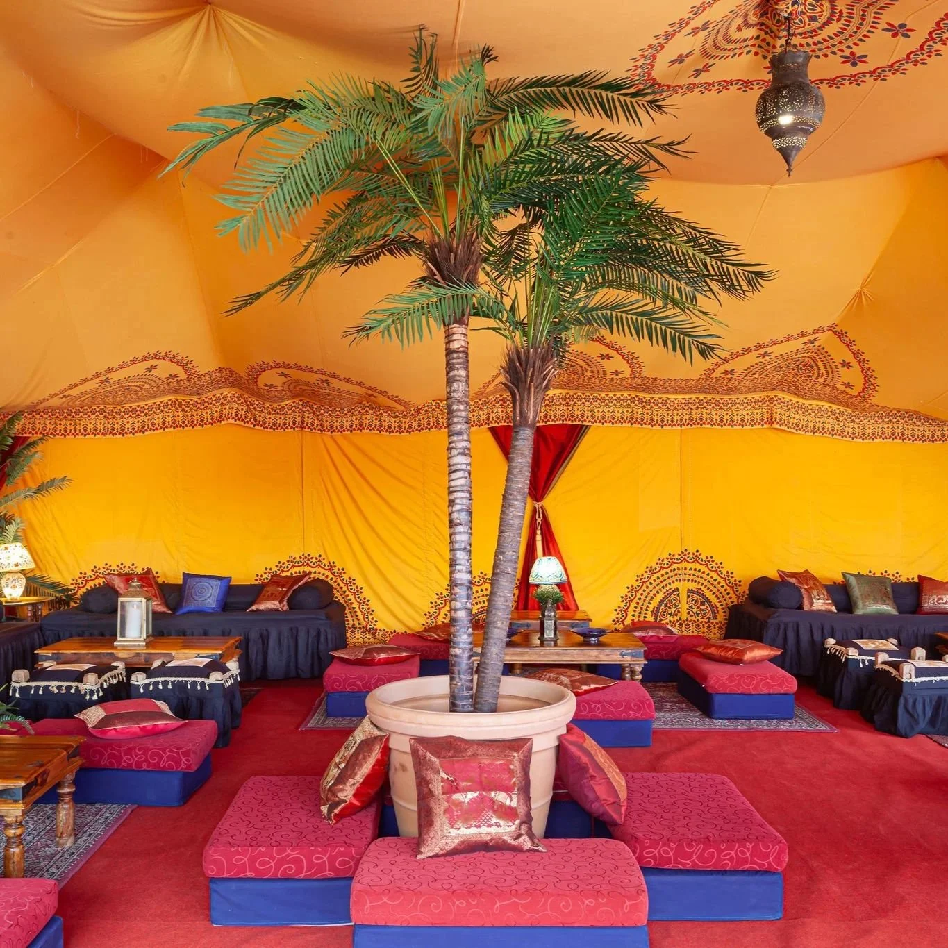 Exotic saffron-yellow marquee interior styled with rich red and blue furnishings, low seating with embroidered cushions, and wooden tables. The vibrant tent design includes palm trees, Moroccan lanterns, and patterned fabric linings, creating a luxur