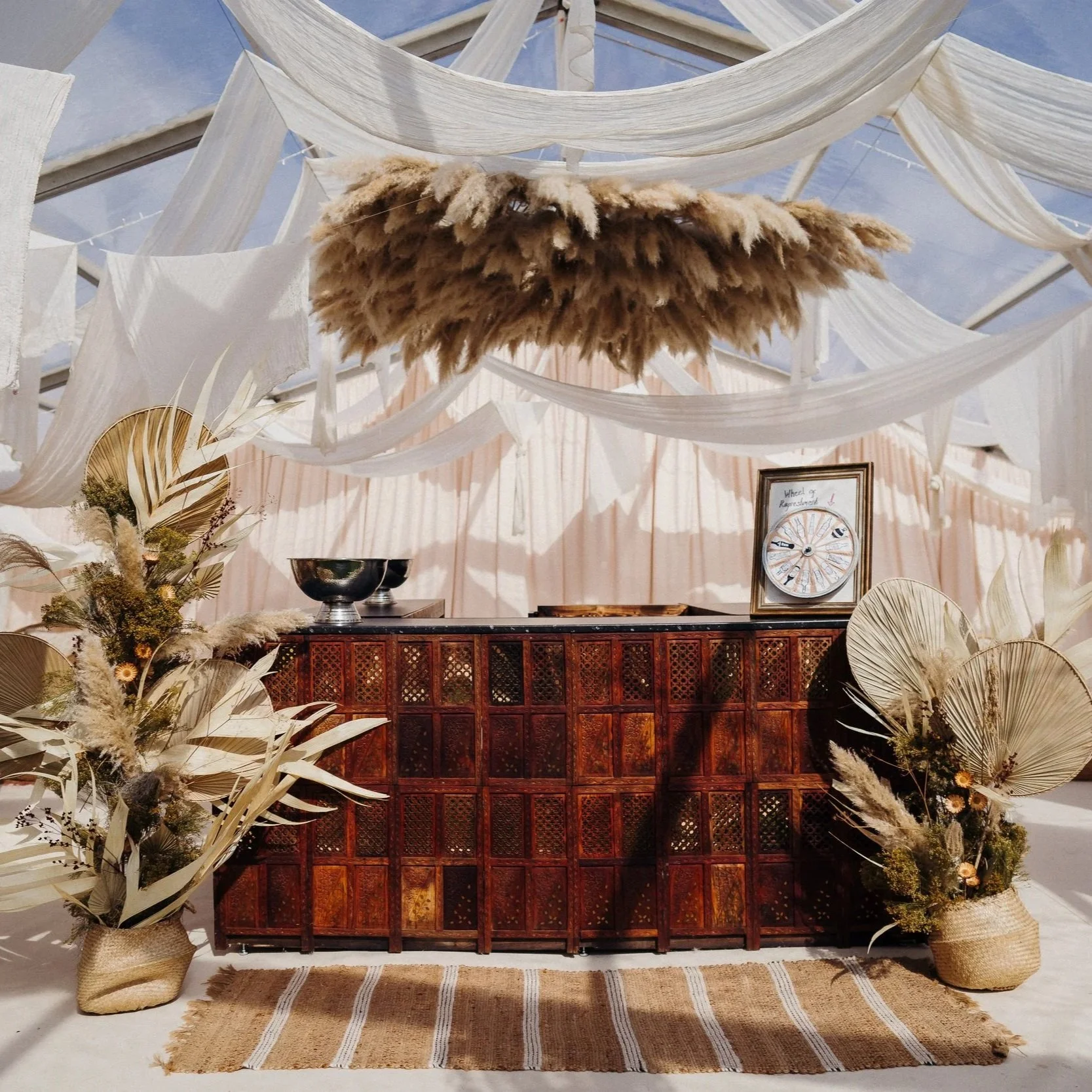 Bohemian-inspired wedding bar inside a clear roof marquee, styled with flowing white drapery, a pampas grass chandelier, and rustic wooden furniture. Dried palm leaves and natural floral arrangements frame the bar, while woven rugs and wicker chairs 