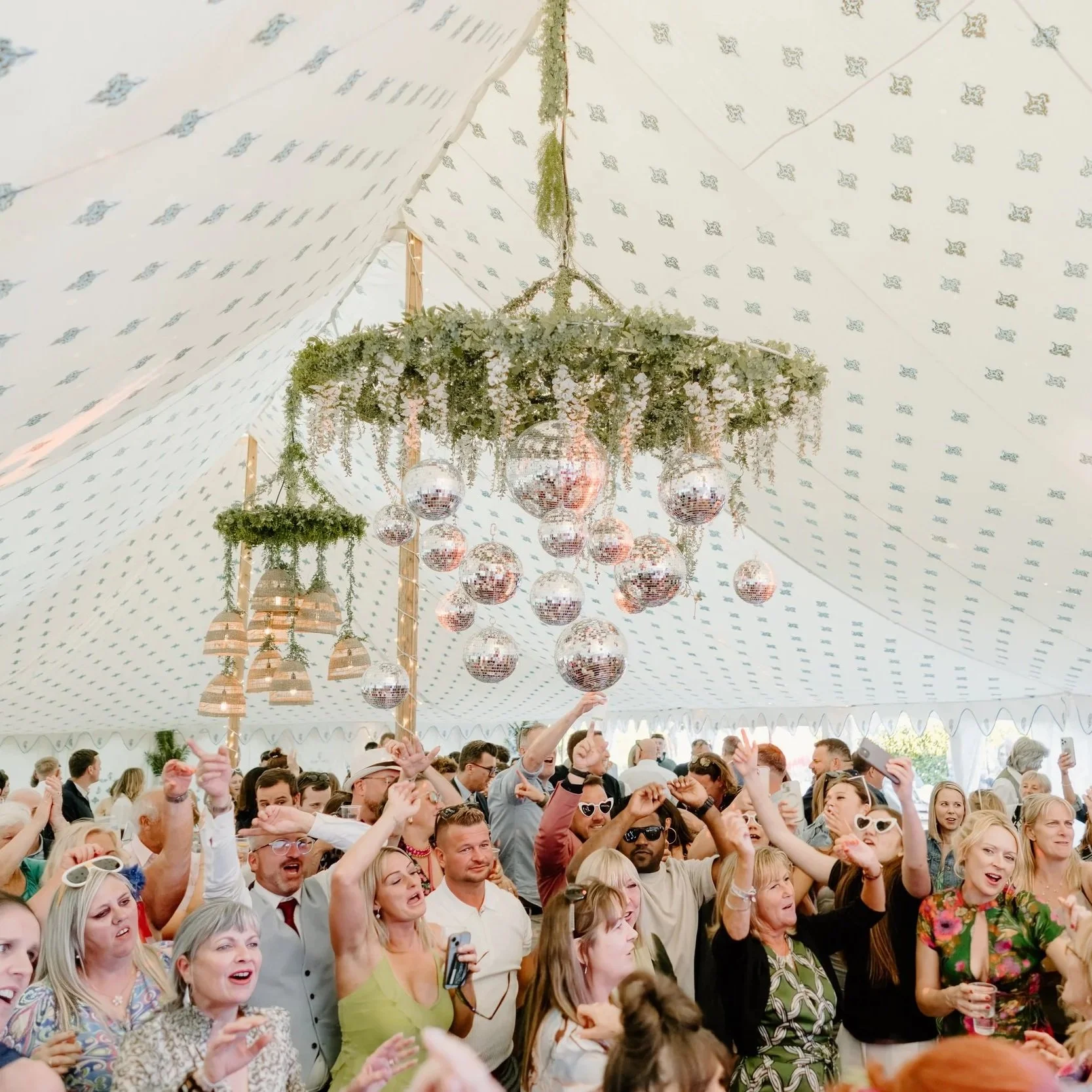 Lively wedding party inside a marquee with patterned cream linings, featuring a floral and greenery chandelier with disco balls hanging above the dance floor. Guests celebrate with dancing, singing, and laughter, creating a vibrant atmosphere. The st