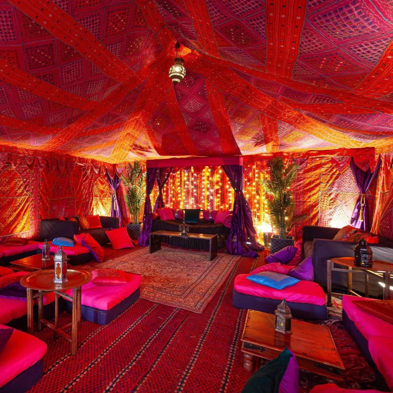 Exotic Indian-inspired marquee interior decorated with vibrant red and purple fabrics, embroidered linings, and low seating with bright cushions. The luxurious lounge area features Moroccan lanterns, patterned rugs, and warm ambient lighting, creatin