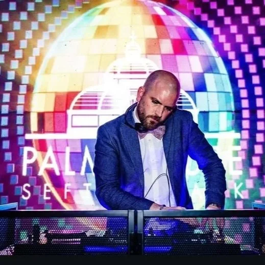 Professional wedding DJ performing live against a vibrant disco ball backdrop, creating an unforgettable party atmosphere. Dressed in a stylish suit and mixing tracks at the booth, this DJ brings energy, music, and lighting together to keep the dance