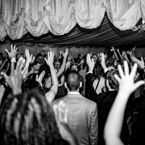 Black and white photo of wedding guests with their hands in the air on a packed dance floor. The lively celebration captures the joy, energy, and atmosphere of a wedding party where music and entertainment keep everyone dancing late into the night.
