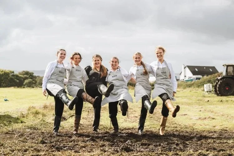The Regis Banqueting catering team smiling and kicking up their wellies outdoors, showing their fun, hardworking spirit during a countryside wedding event. Dressed in matching aprons, the team reflects professionalism, teamwork, and dedication to pro