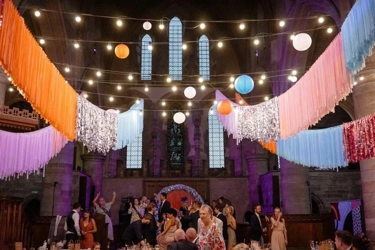 A vibrant wedding reception styled by Streamadelica Event Styling, featuring colourful hanging fringe garlands in orange, pink, blue, and silver beneath the grand arches of a historic venue. Paper lanterns and string lights add a whimsical glow above