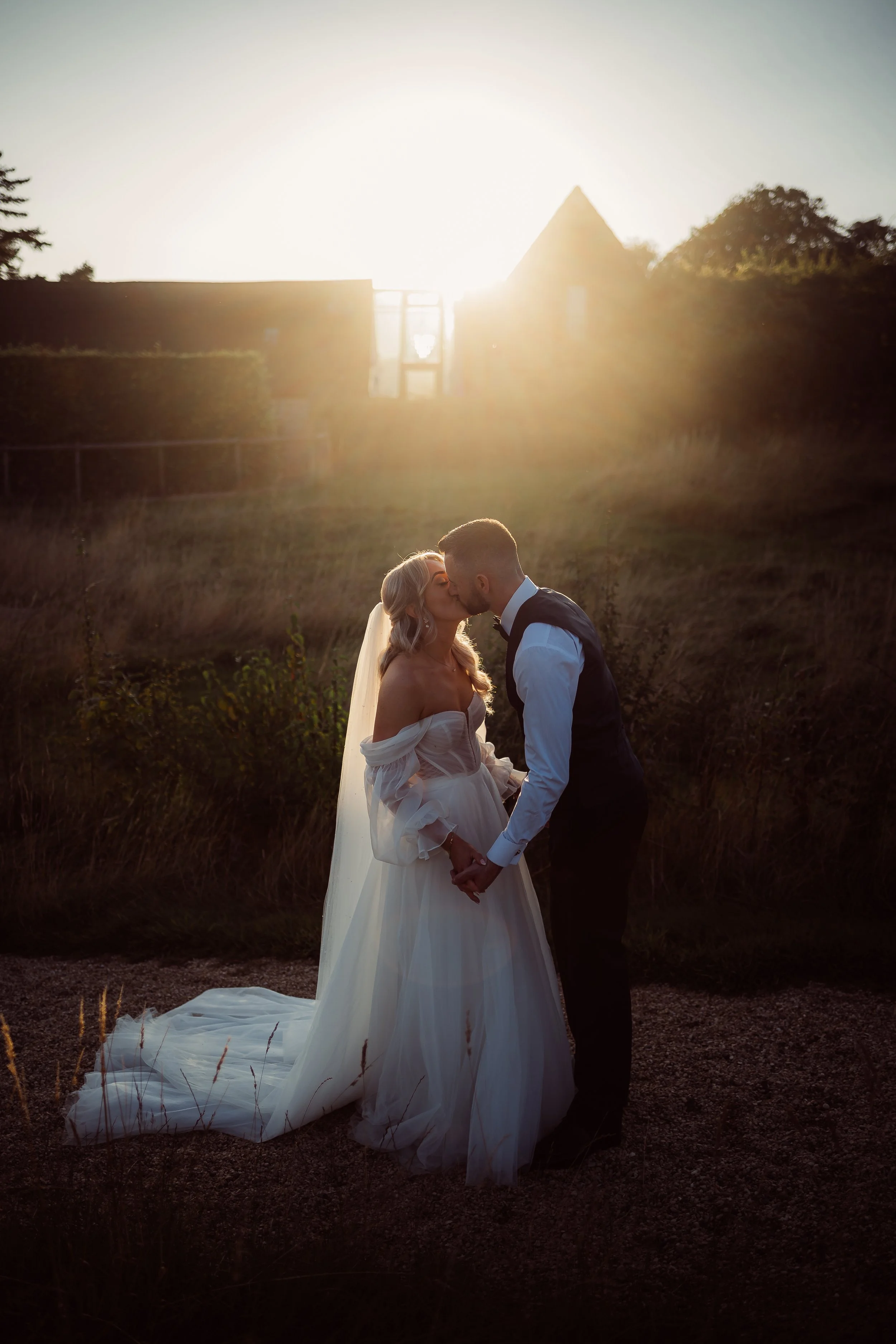 A romantic golden-hour wedding portrait by Gemma Gaskins Photography, featuring a couple sharing a kiss in soft sunset light. The bride’s flowing veil and off-the-shoulder gown glow in the warm backlight while the groom holds her hands, creating a dr