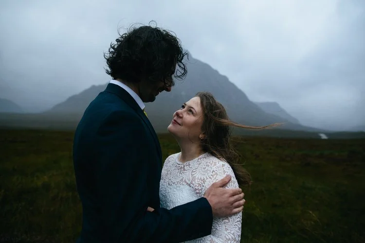 An intimate outdoor wedding portrait captured by Mirrorbox Photography, featuring a couple embracing in the dramatic landscape of the Scottish Highlands. The bride, wearing a long-sleeved lace dress, looks up at her partner with a soft, windswept smi