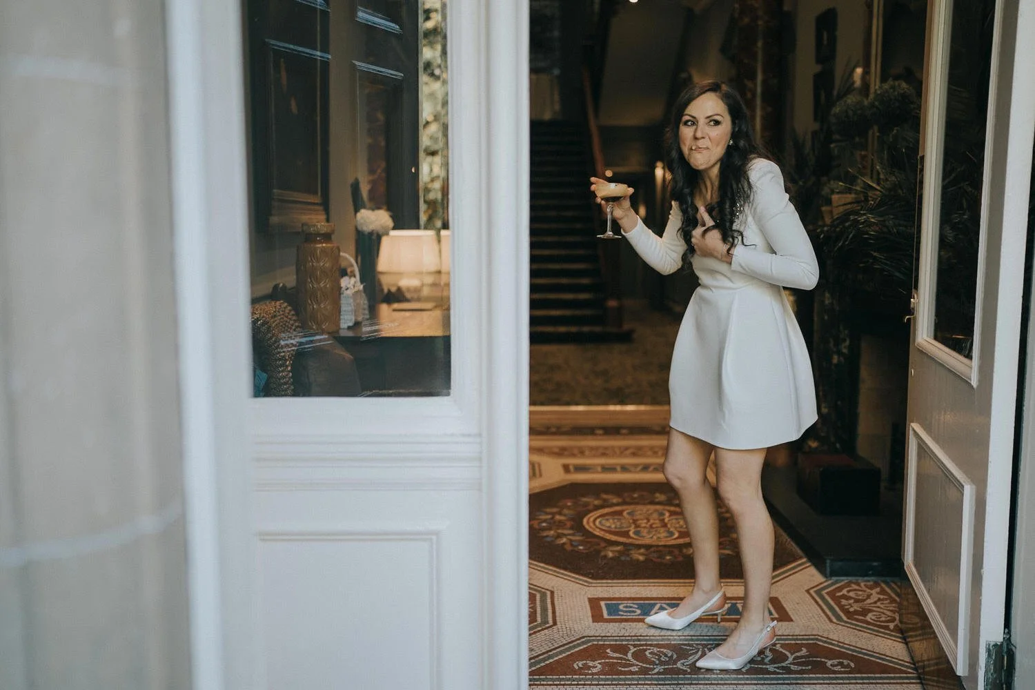 A candid wedding moment captured by Good Luck Wolf Photography showing a bride in a short white wedding dress and heels laughing as she steps through a grand doorway holding a cocktail, with patterned tiles and a sweeping staircase behind her, full o