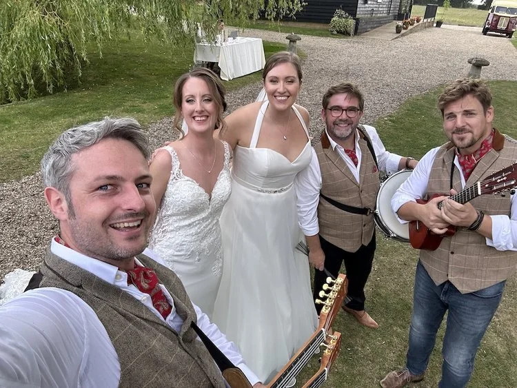 The Ukes of Hazard pose for a cheerful selfie with two brides at an outdoor wedding, dressed in matching tweed waistcoats, white shirts, and red neckerchiefs. With guitars and a banjo in hand, the acoustic band radiate warmth and personality, perfect