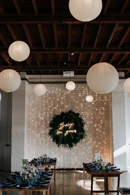 Modern industrial wedding setup featuring a lush greenery wreath by Catherine Short Florals displayed with a neon sign against a backdrop of fairy lights. Paired with long wooden tables styled with soft floral arrangements and elegant place settings,