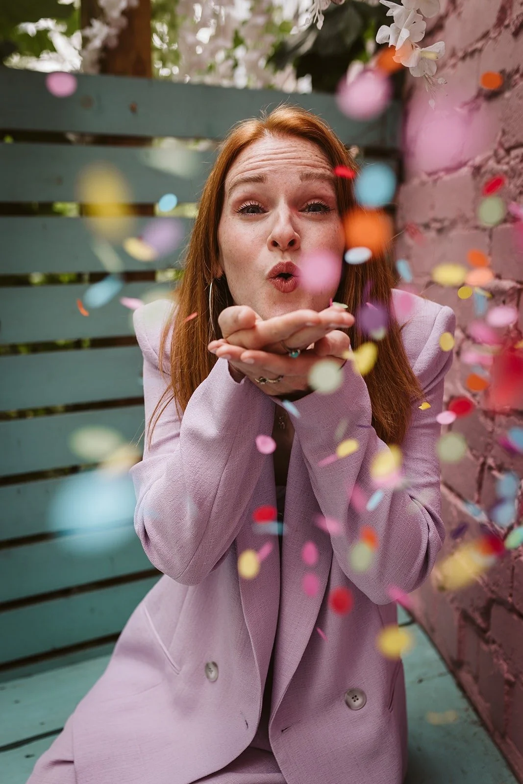 Wedding celebrant Meg Senior wearing a lilac suit joyfully blows colorful confetti toward the camera. The playful moment is captured against a pastel backdrop of teal wood and pink brick, perfectly reflecting her fun, modern, and creative celebrant s