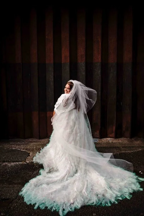 A cinematic bridal portrait captured by Amy Faith Photography, featuring a bride standing against a dramatically textured industrial backdrop. She wears an ethereal, feather-textured wedding gown with an extravagant train that pools around her in sof