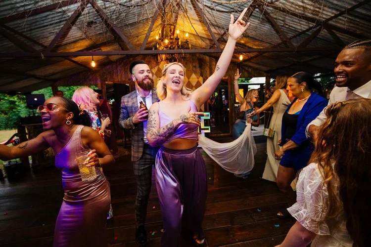 A vibrant wedding reception dance floor scene with guests celebrating under warm, rustic lighting. A bridesmaid in a shiny purple two-piece outfit leads the group with her arm raised, singing and dancing with a drink in hand. Other guests around her 