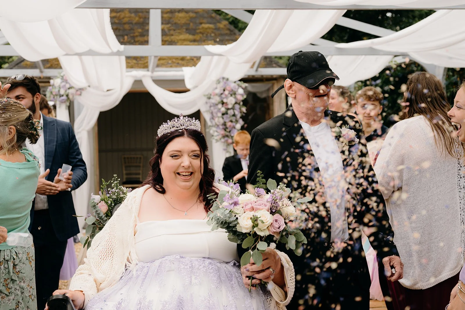 A bride in a wheelchair and her groom are being covered in dried flower confetti. They both look very happy.