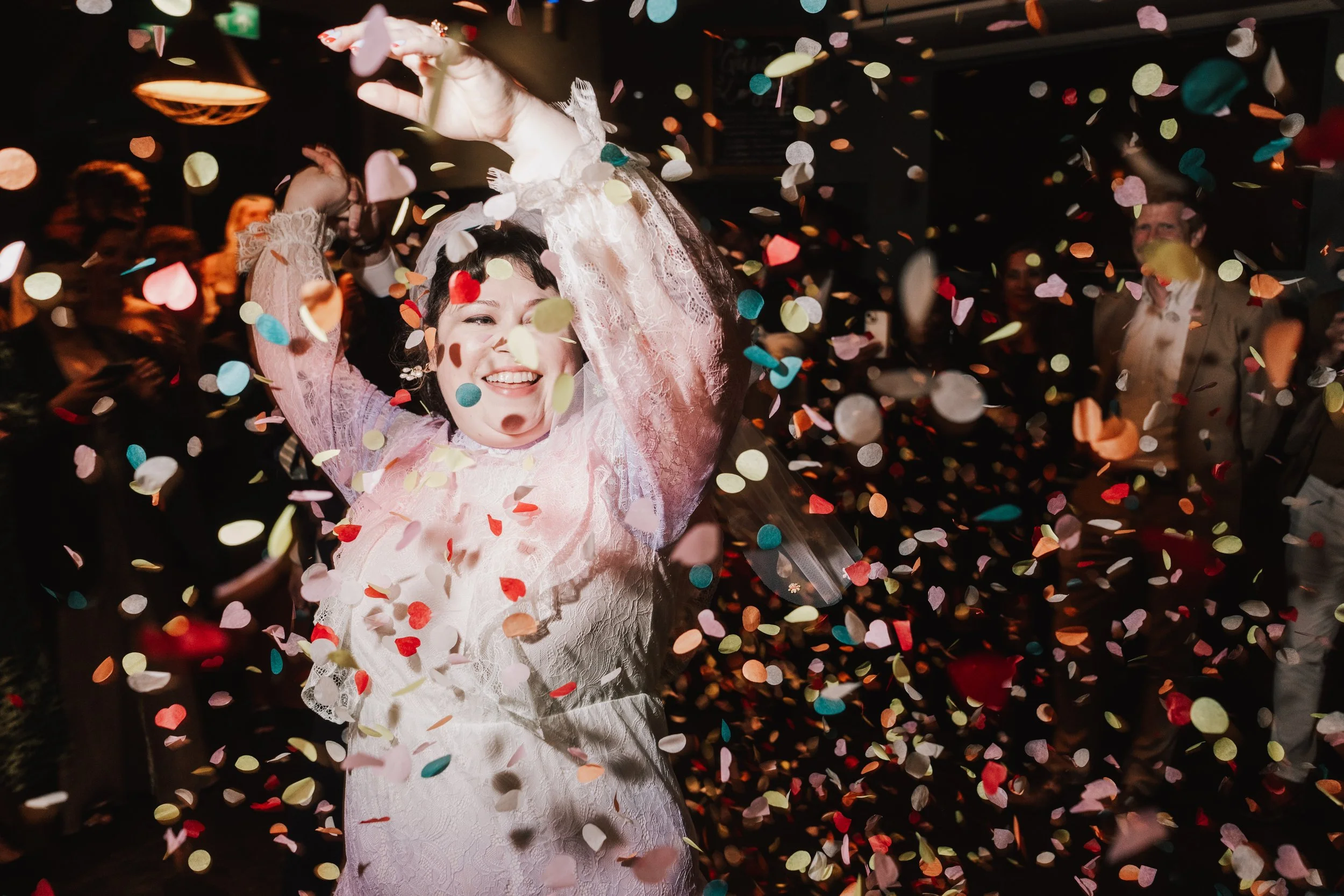 A joyful bride dances through a vibrant shower of multicoloured confetti, her lace dress glowing under the warm reception lights as guests cheer in the background. Her laughter and raised arms capture the pure energy and celebration of the moment, cr