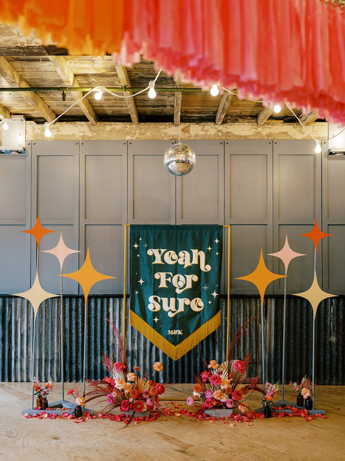 Retro-inspired wedding backdrop by AMW Events featuring a green and gold “Yeah For Sure” banner, disco ball, and vibrant red and pink floral arrangements with playful starburst cut-outs under warm string lights.