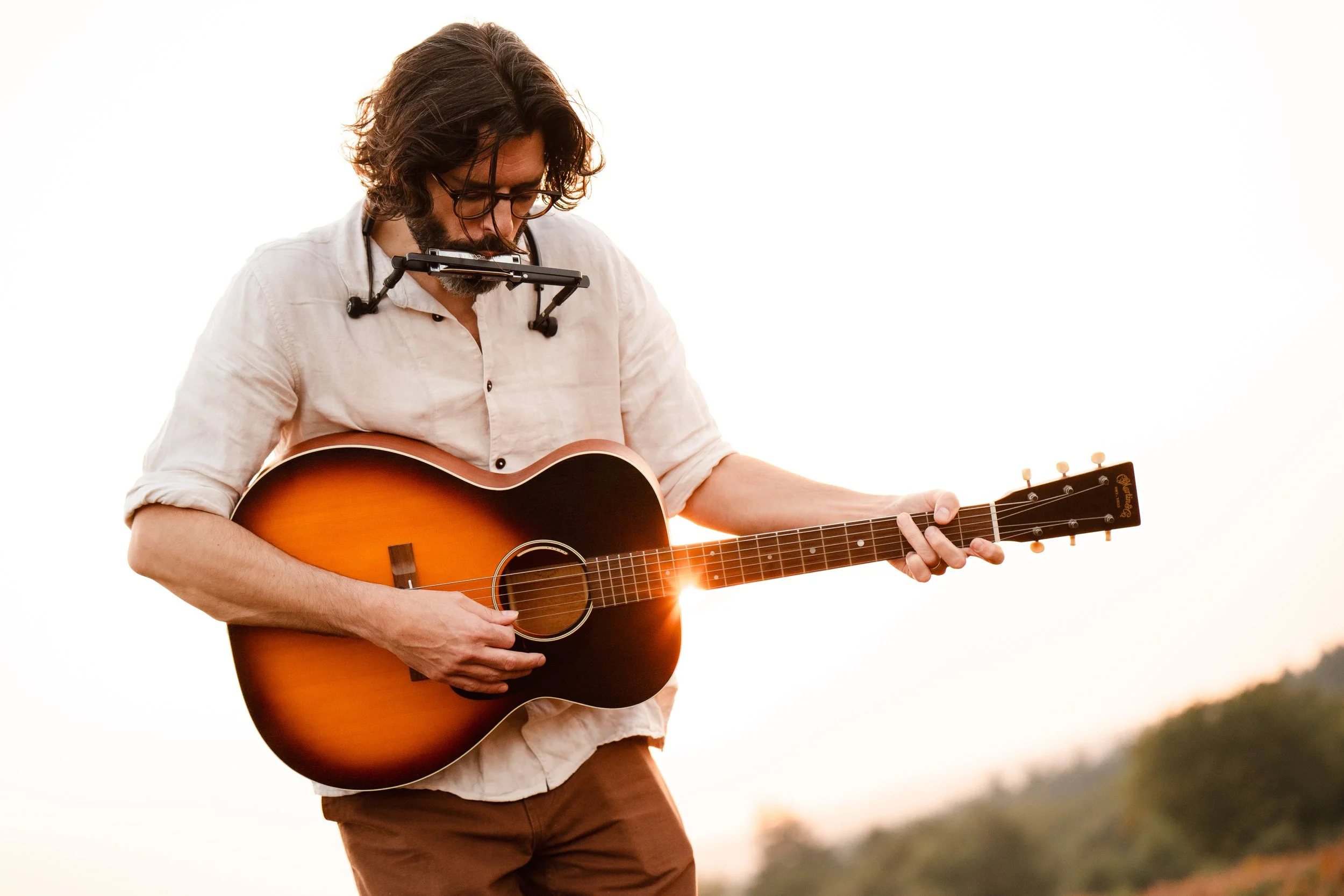 Thomas J Wilman performs outdoors at sunset, playing acoustic guitar with a harmonica for a relaxed, heartfelt wedding set. His warm, soulful style makes him a sought-after acoustic wedding singer and guitarist across the UK. Clarkie Photography.