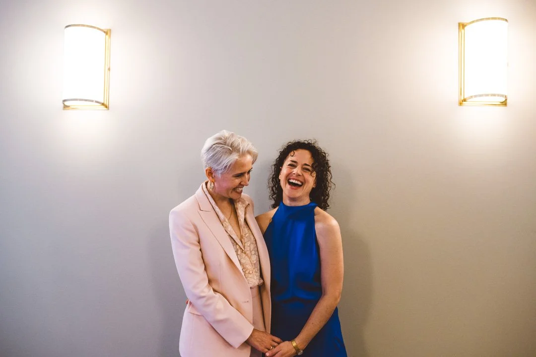 A joyful and intimate portrait captured by Barbara K Photography, featuring a couple sharing a relaxed moment of laughter against a minimalist wall illuminated by warm sconces. One partner wears a tailored blush suit while the other wears a vibrant b
