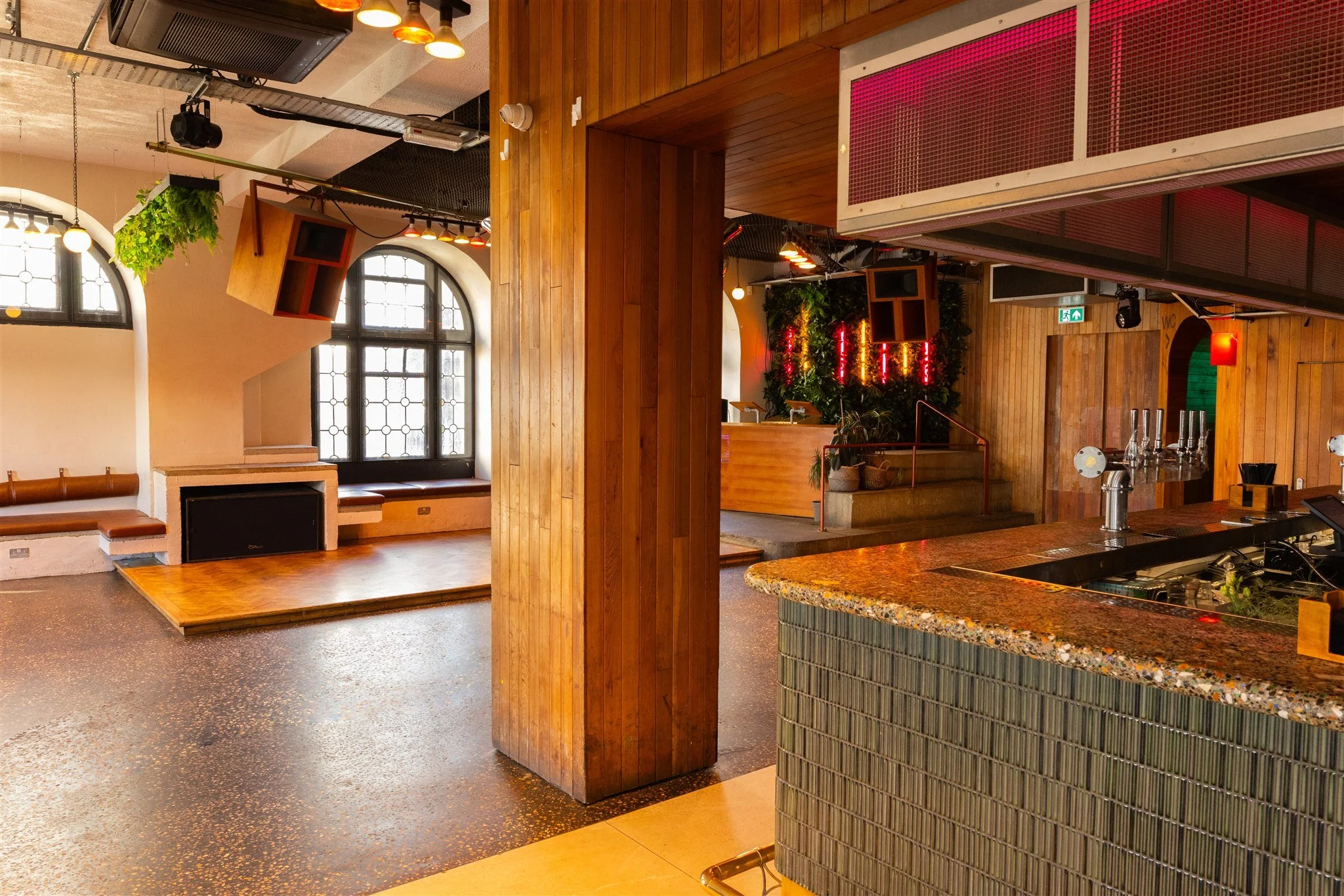 The bar and lounge area inside Pier Court Brighton wedding venue showcasing its unique blend of natural wood finishes, terrazzo-style bar top, arched windows, indoor greenery, and ambient neon lighting. This flexible celebration space offers cosy bui