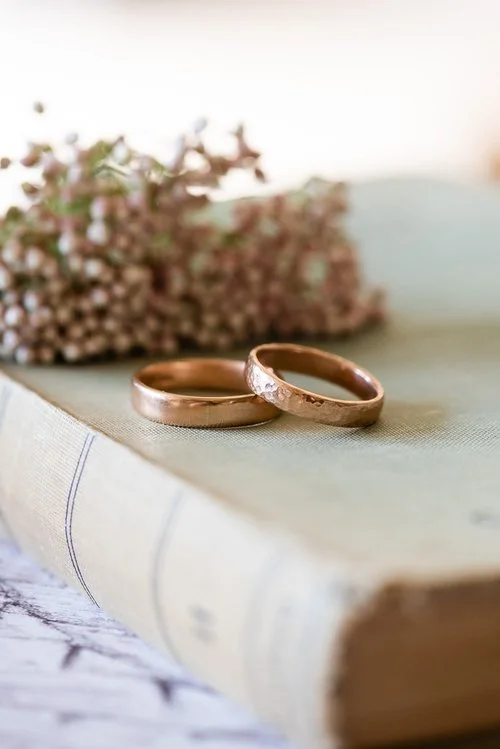 Pair of handcrafted rose gold wedding bands by Nikki Stark Jewellery, resting on a vintage book with floral detail. Made in the UK, these elegant hammered rings are ideal for couples seeking timeless, bespoke wedding jewellery.