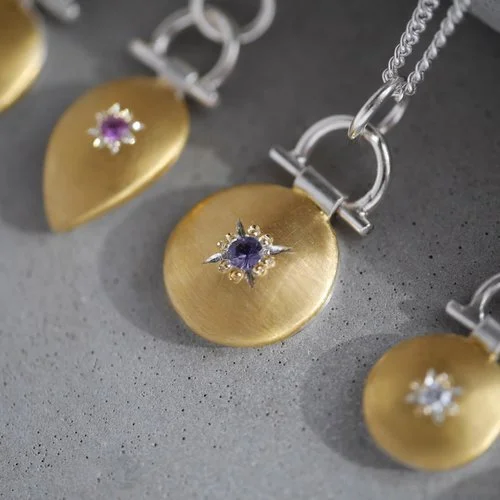 Handcrafted Natalie Vernazza pendant featuring a brushed gold disc set with a purple-blue spinel gemstone, suspended from a silver chain, showcasing modern artisan wedding jewellery with refined texture and elegant contrast.