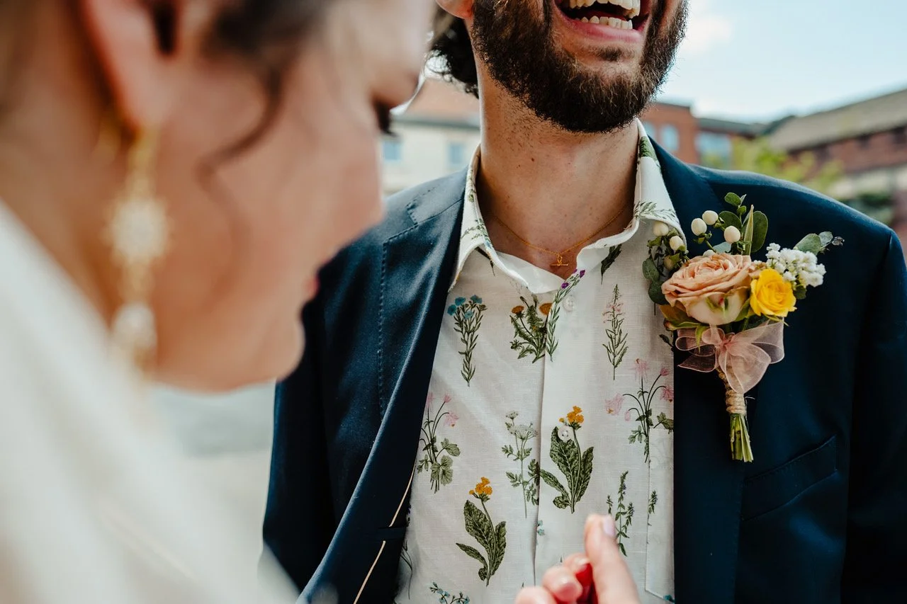 A close crop captures the joyful warmth between two people as the focus falls on a laughing man in a navy jacket paired with a botanical print shirt and a pastel boutonniere, while the softly blurred profile of his partner and her gold earrings frame