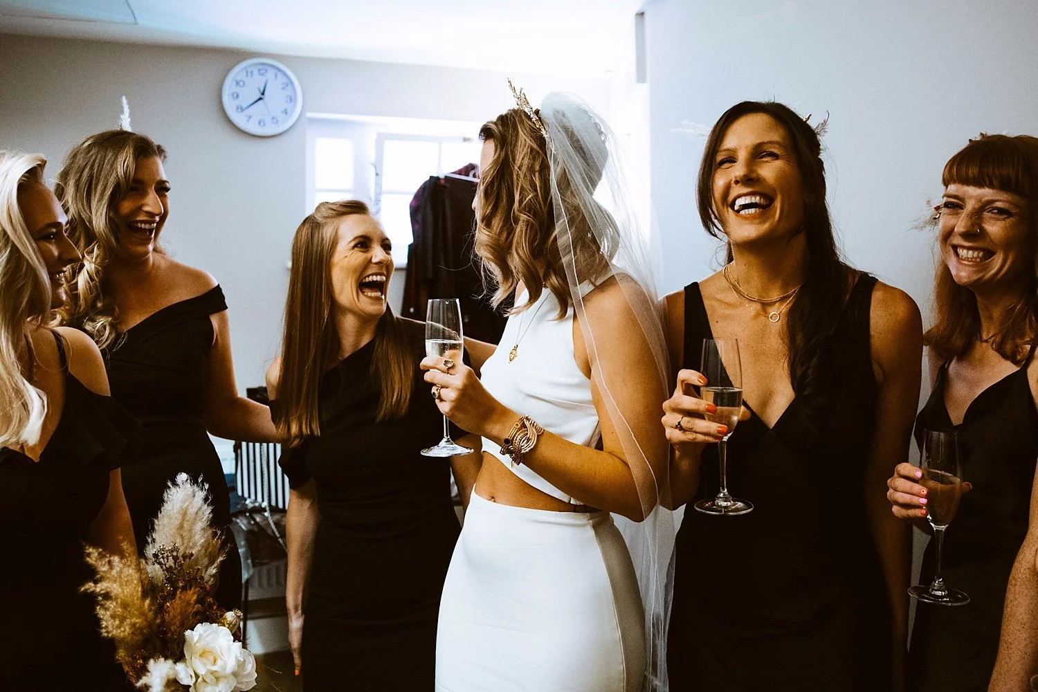 A room buzzing with pre-wedding excitement as the bride stands at the centre of her crew, all of them mid-laugh with champagne glasses in hand. She wears a sleek white two-piece with a modern cut-out waist and a delicate veil, framed by friends in ch