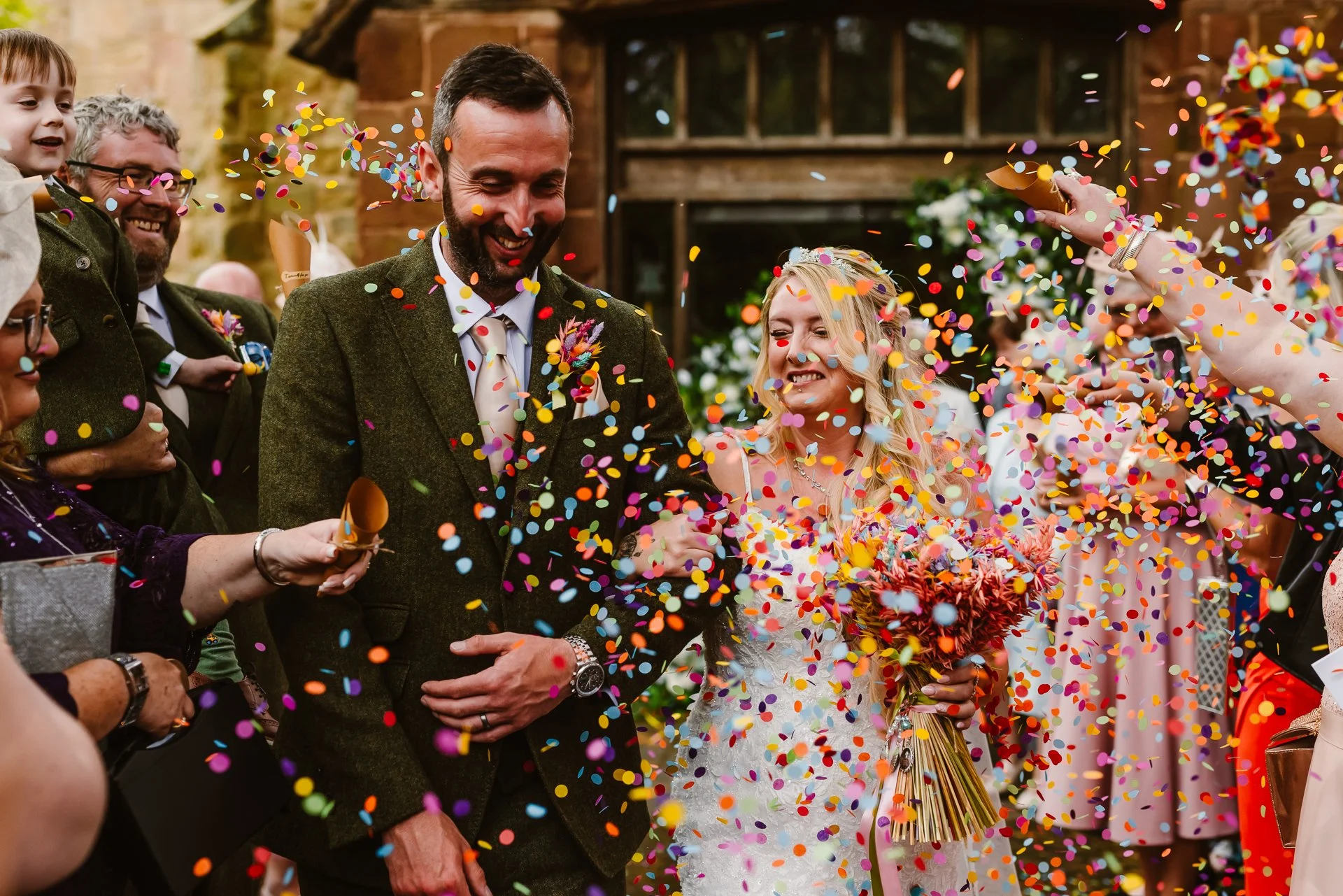 A joyful wedding confetti moment captured by Bluebell Photo Studio, showing a newly married couple walking hand in hand through a colourful shower of petals outside their rustic UK venue. The bride smiles brightly with her vibrant bouquet while guest