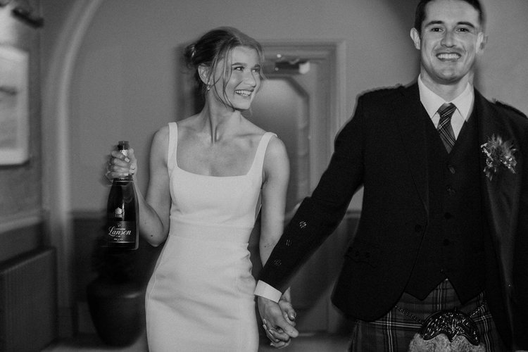 A joyful black-and-white moment captured by Mirrorbox Photography, showing a newly married couple walking hand-in-hand through their wedding venue. The bride, wearing a sleek modern gown, smiles while holding a bottle of champagne, perfectly conveyin