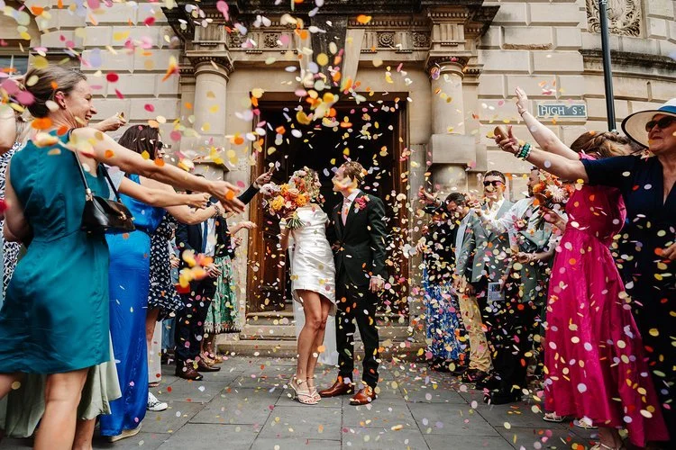 A newly married couple share a kiss beneath a vibrant shower of multicoloured confetti as they step out of an ornate building onto a bustling high street. Friends and family line the path on both sides, cheering and throwing handfuls of petals while 