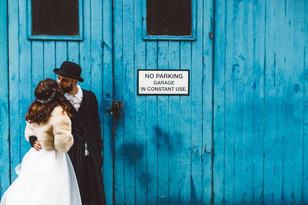 A creative and stylish wedding portrait captured by Barbara K Photography, featuring a couple sharing a quiet kiss in front of a bold blue garage door. The bride wears a classic white gown paired with a vintage inspired fur jacket and a beaded headpi