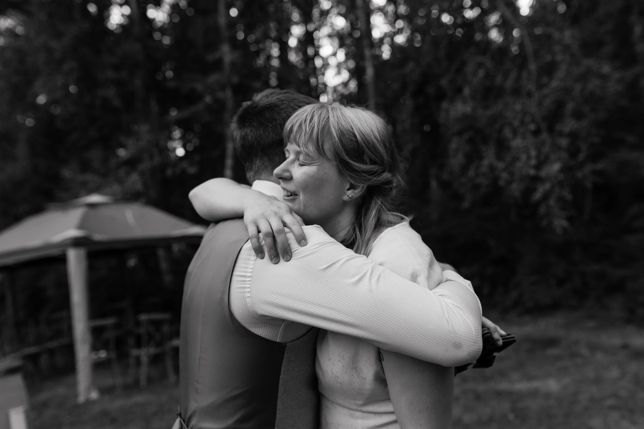 A tender black-and-white wedding moment captured by Laura Wilson Photography, showing a couple sharing a heartfelt embrace outdoors. The bride closes her eyes with a peaceful, emotional expression as she wraps her arms around her partner, whose back 