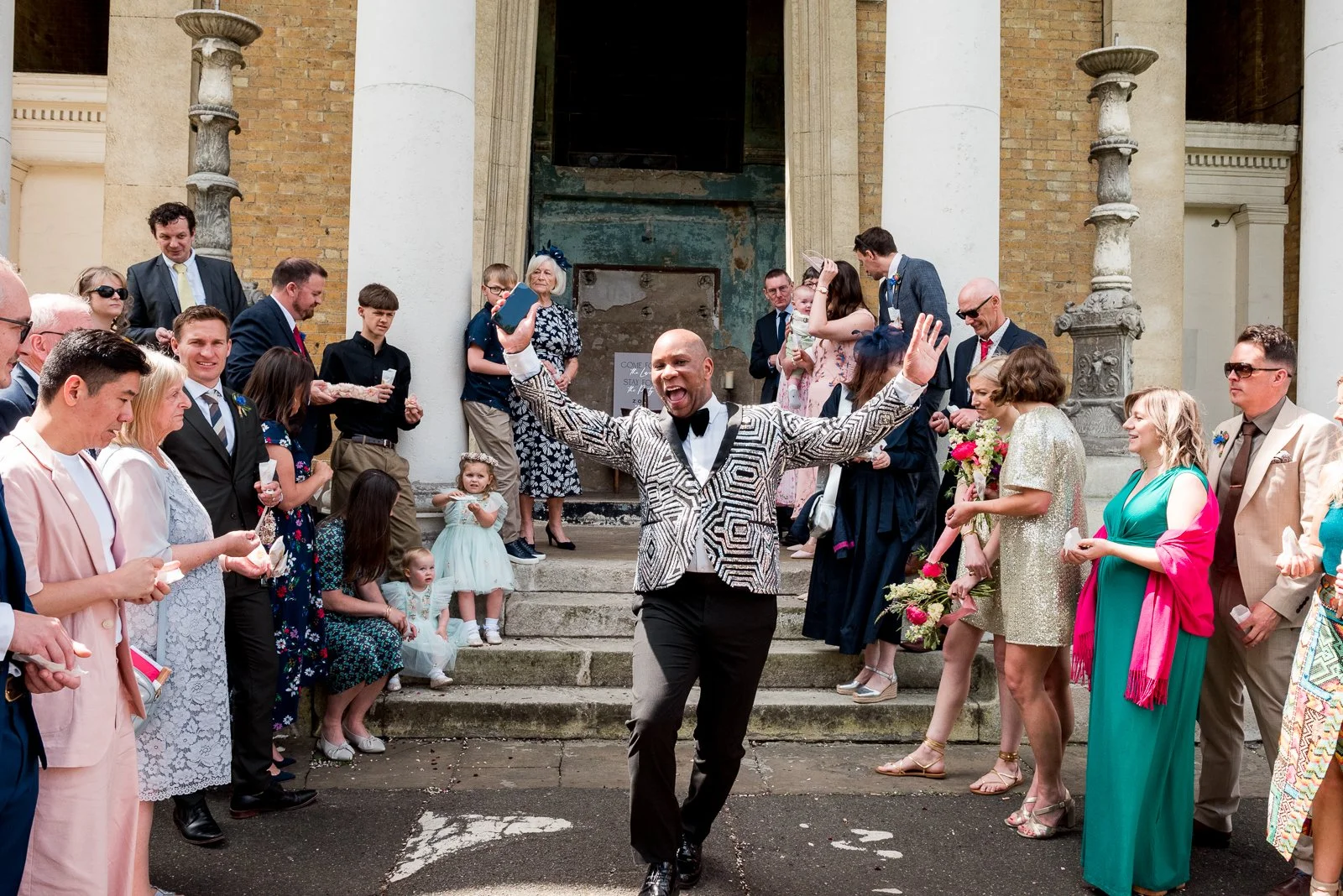 Dave Octave Celebrant in a black and white zebra print blazer, standing in front of a building with his hands in the air, surrounded by members of the wedding congregation