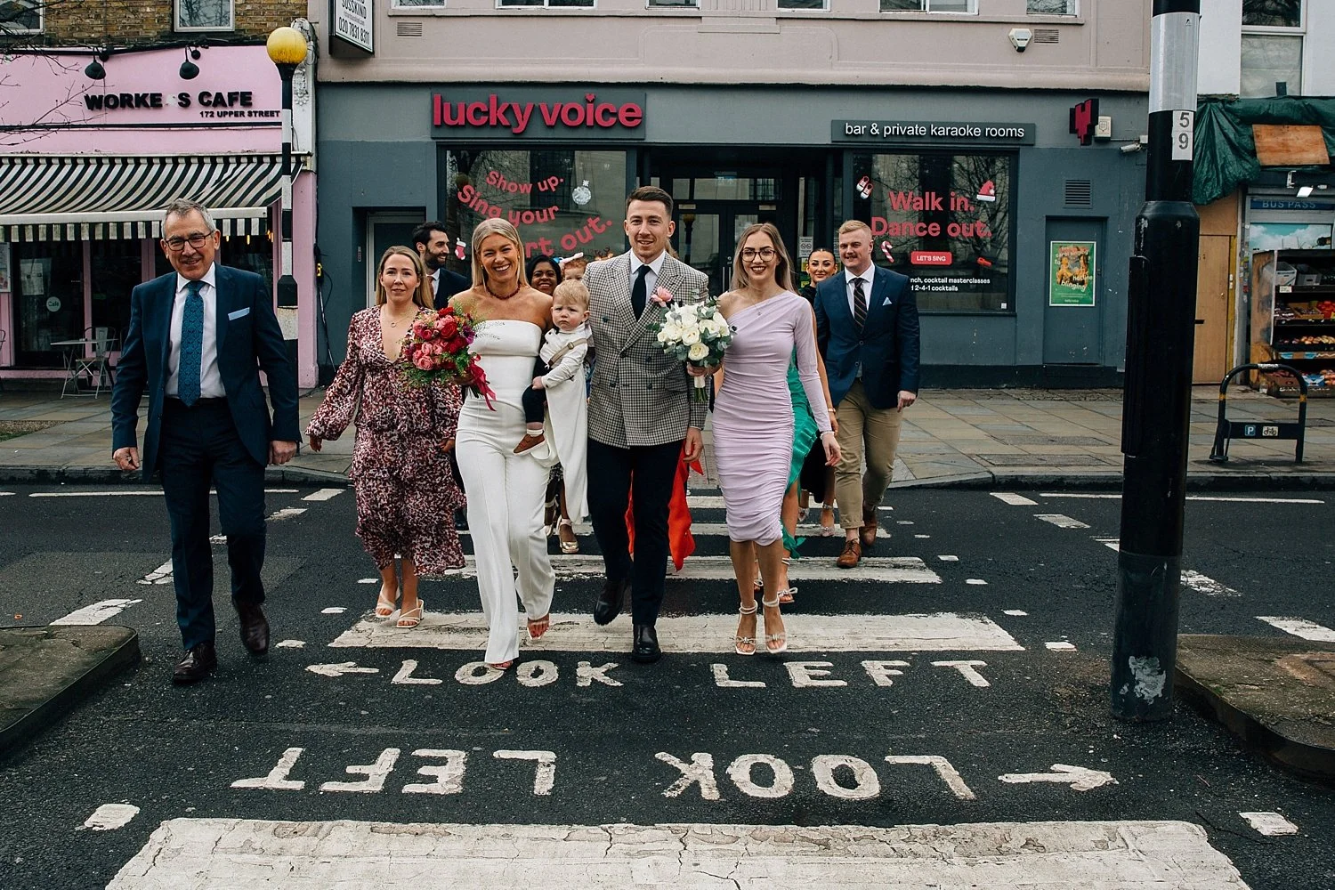 A lively, cinematic moment as the wedding party takes over a London zebra crossing with total swagger and joy. The couple leads the pack - she’s radiant in a chic white jumpsuit, bouquet bursting with bold pinks; he’s sharp in a checked blazer, carry
