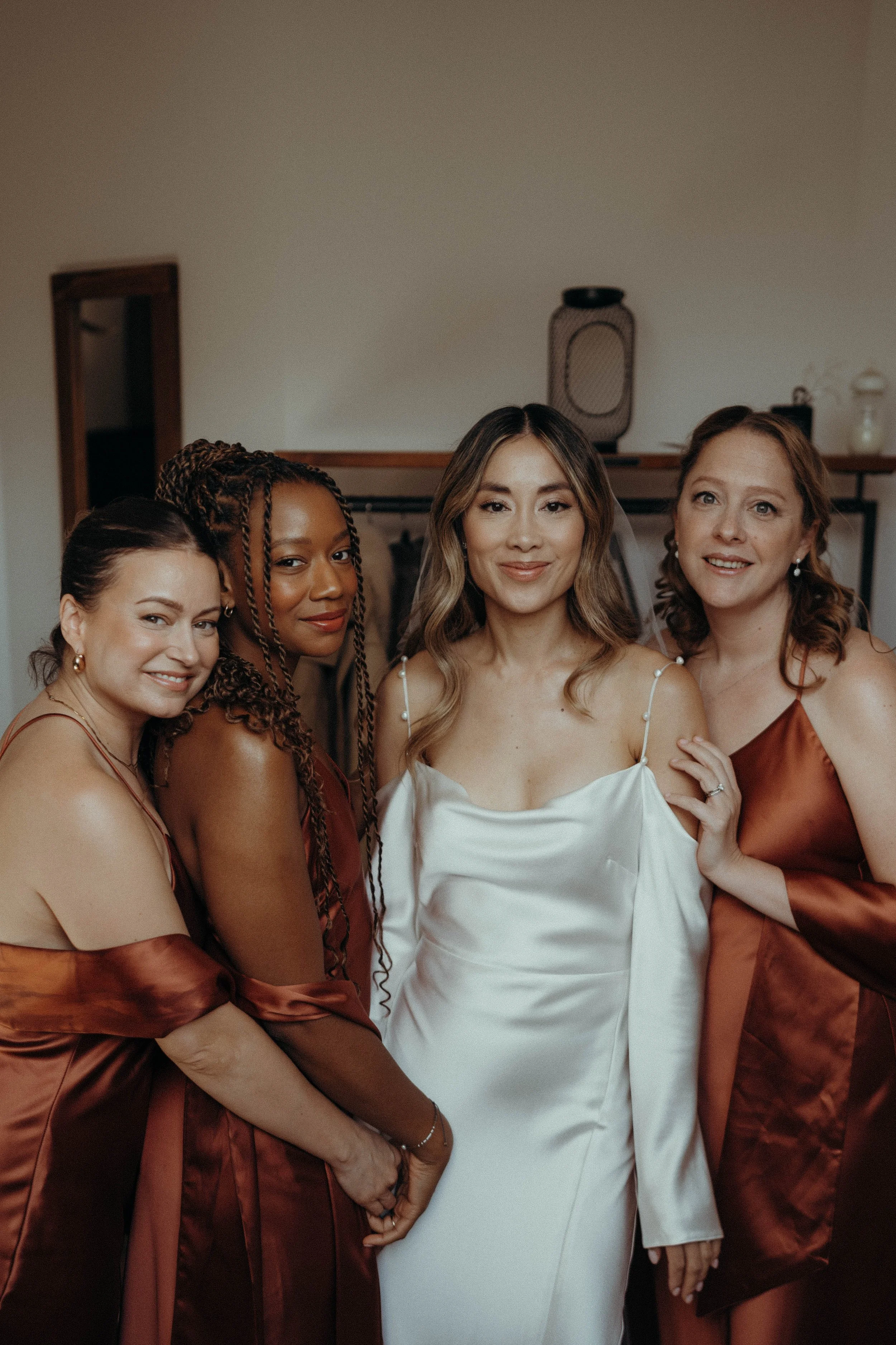 A timeless and modern look by Rewritten Bridal Wear, featuring a sleek satin bridal gown with elegant off-the-shoulder sleeves and a soft cowl neckline. The bride’s effortless style is complemented by her bridesmaids in rich copper-toned satin dresse