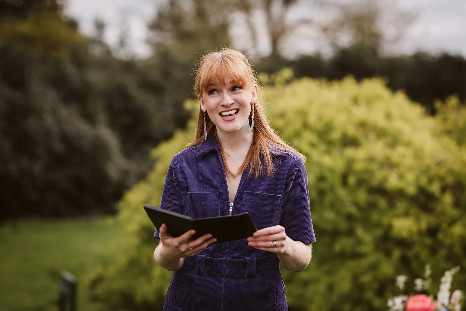 This image shows wedding celebrant Meg Senior speaking outdoors, holding an open book or folder. She’s dressed in a deep purple outfit, standing against a lush, green background with trees and bushes. The setting and her expression suggest she’s lead