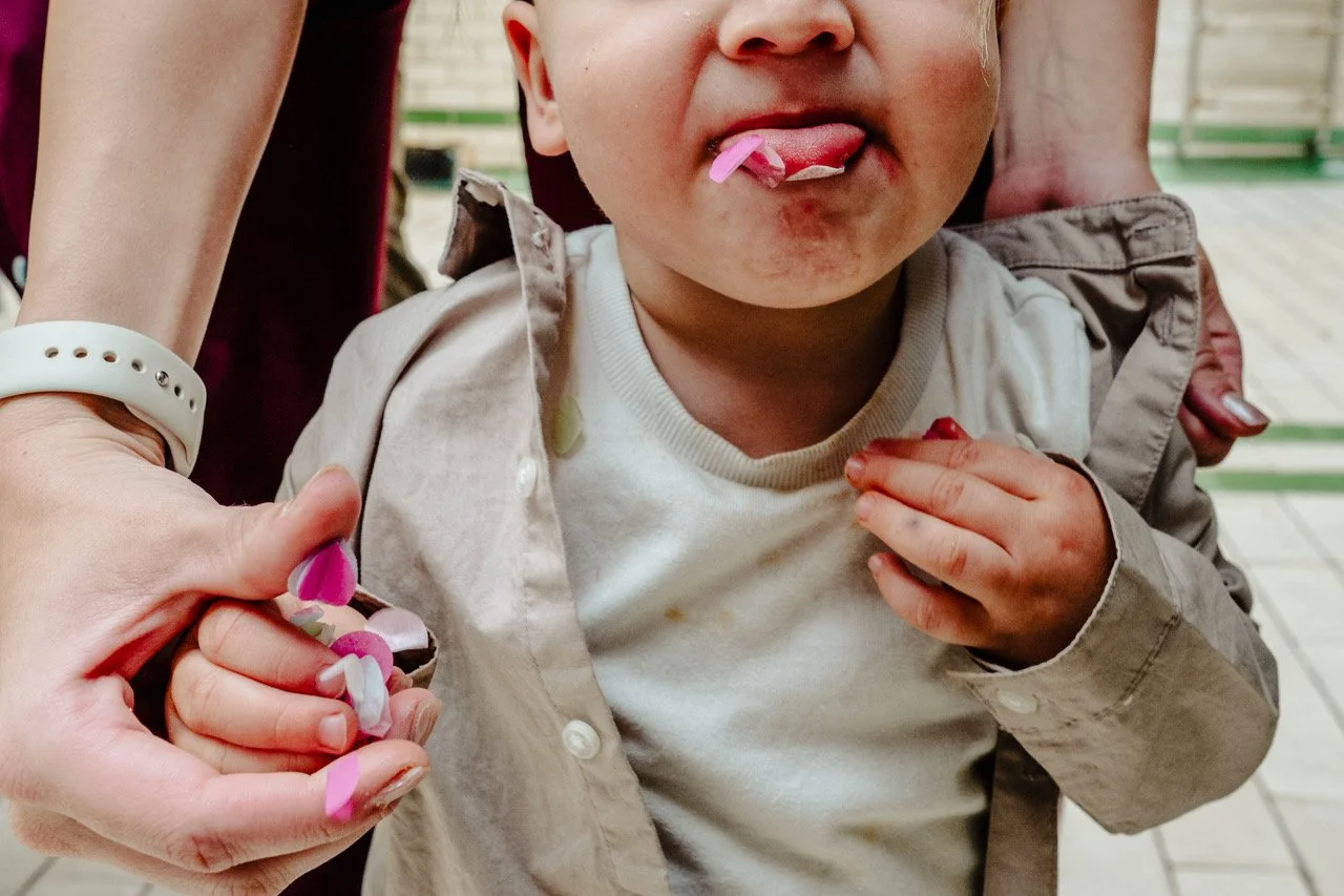 A toddler stands clutching an adult’s hand with fingers full of colourful confetti, cheeks flushed and shirt slightly stained, mischievously sticking out their tongue with pink petals stuck to it while someone steadies them from behind in a bright ti