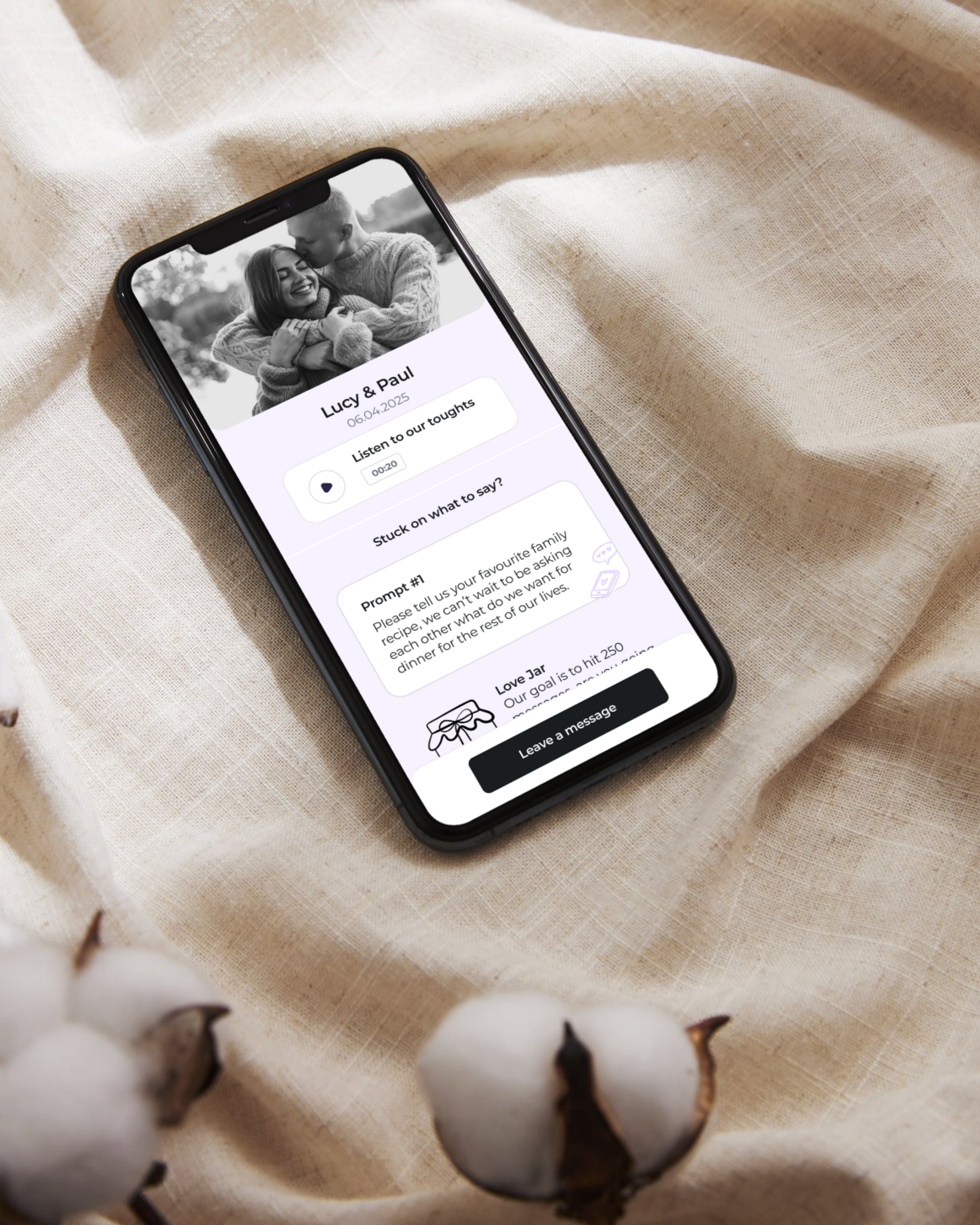 A smartphone displaying the VOXU Audio Guestbook interface for Lucy & Paul’s wedding on 6th April 2025, resting on soft linen fabric with cotton flowers. The elegant design shows prompts for guests to record personal voice notes, creating meaningful 