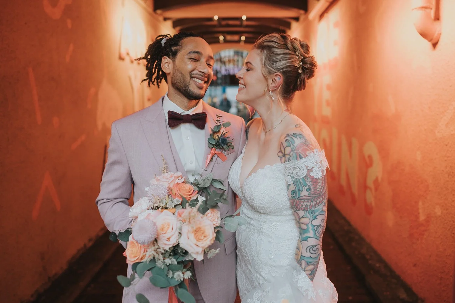 An intimate wedding portrait by Good Luck Wolf Photography showing a tattooed bride in an off-the-shoulder lace gown and a groom in a blush suit sharing a laugh in a warm, orange-lit passageway, capturing relaxed, documentary-style wedding photograph