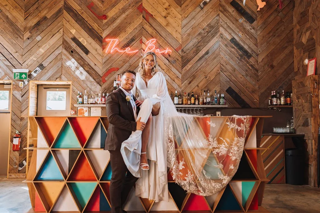 A couple stands at a geometric bar inside a wood panelled room as the groom smiles and supports the bride who is perched on the counter in a flowing white gown with a long embroidered veil that drapes over the colourful triangular shelves while a neo