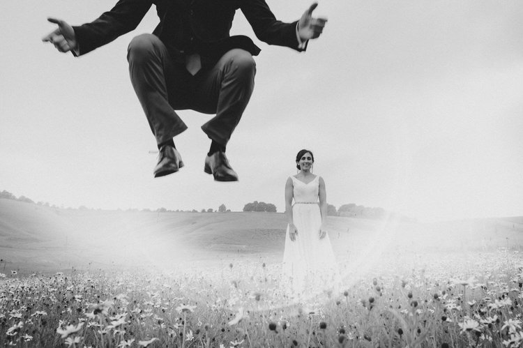 A creative and playful wedding portrait captured by Matt Fox Photography, showing the groom mid jump in the foreground while the bride stands smiling in a wildflower meadow behind him. The black and white edit adds drama and contrast, making the dyna