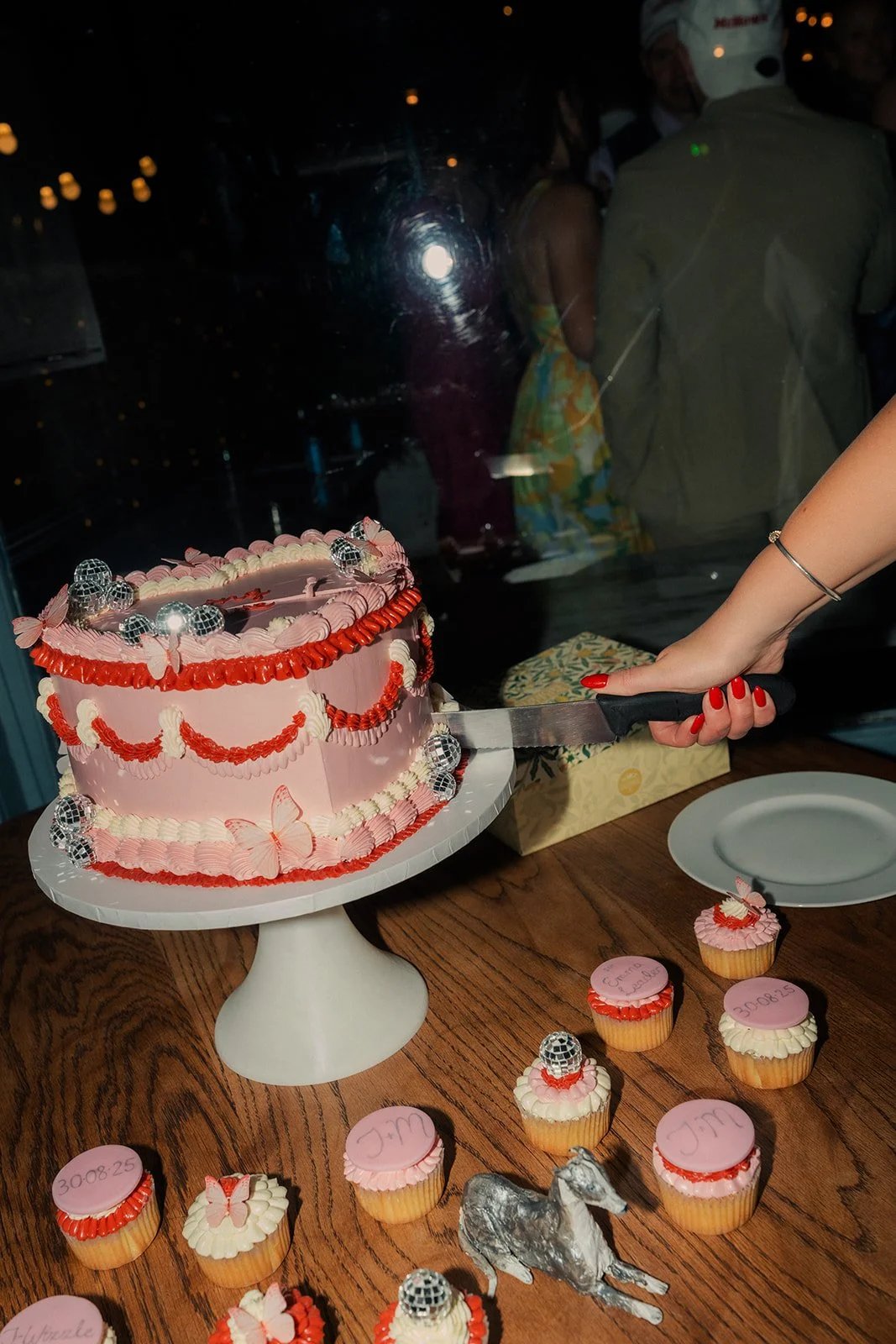 Playful, retro-inspired wedding cake by The Cake Hun featuring vintage piping, soft pink buttercream, red detailing and disco ball accents, styled with matching cupcakes.