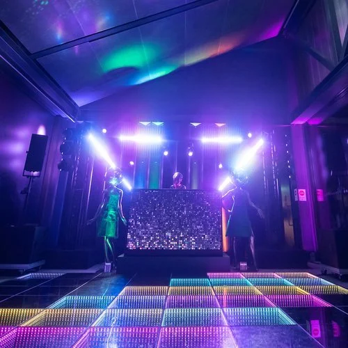 My Colour is Gold sequin DJ booth with dancers in disco ball helmets, lit with vibrant purple and blue lights for a dazzling wedding or party setup.