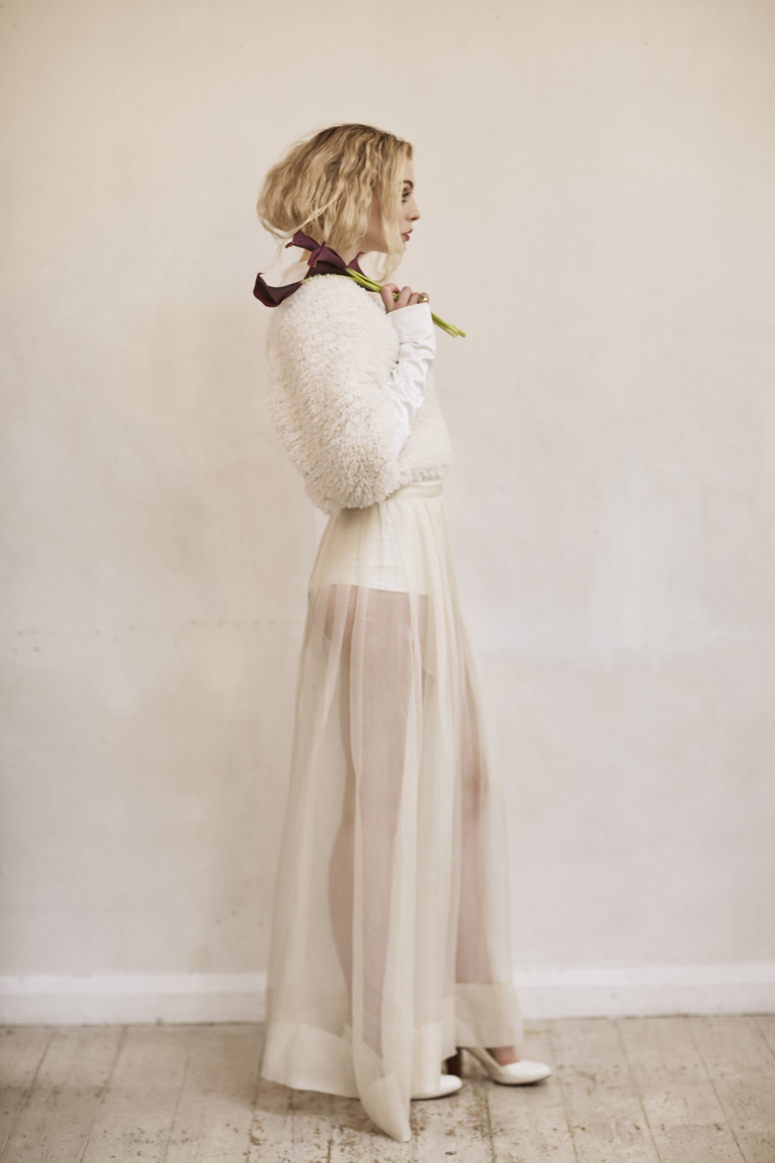 Shown in profile against a pale, minimal backdrop, the look layers soft textures and transparency. A cropped, fluffy jacket sits over a sheer top, while a long, translucent skirt reveals clean lines beneath. A ribbon tied at the neck and a single gre