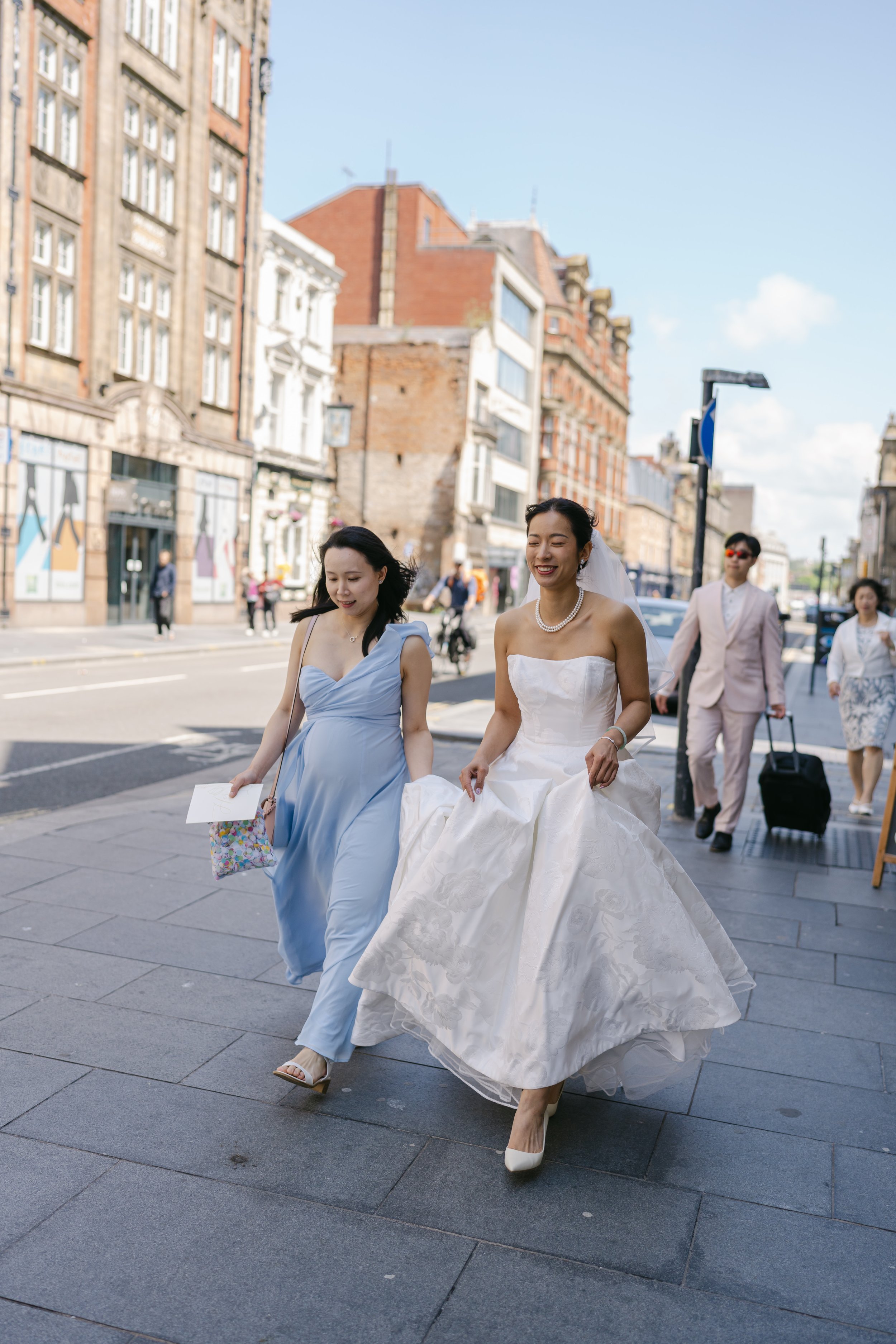 A bride walks through a lively city street with her bridesmaid, laughing together as she lifts her gown, beautifully captured by Christy Photography. The mix of historic buildings, soft sunlight and everyday urban activity creates a modern, authentic