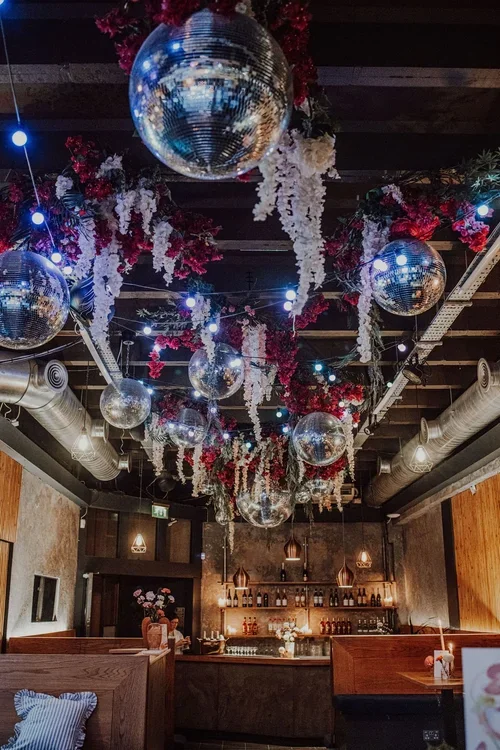 A stylish cocktail bar setup inside Colours Hoxton in Shoreditch, London, featuring a ceiling adorned with sparkling disco balls and cascading floral installations. The warm lighting and industrial-chic decor create a glamorous and modern setting for
