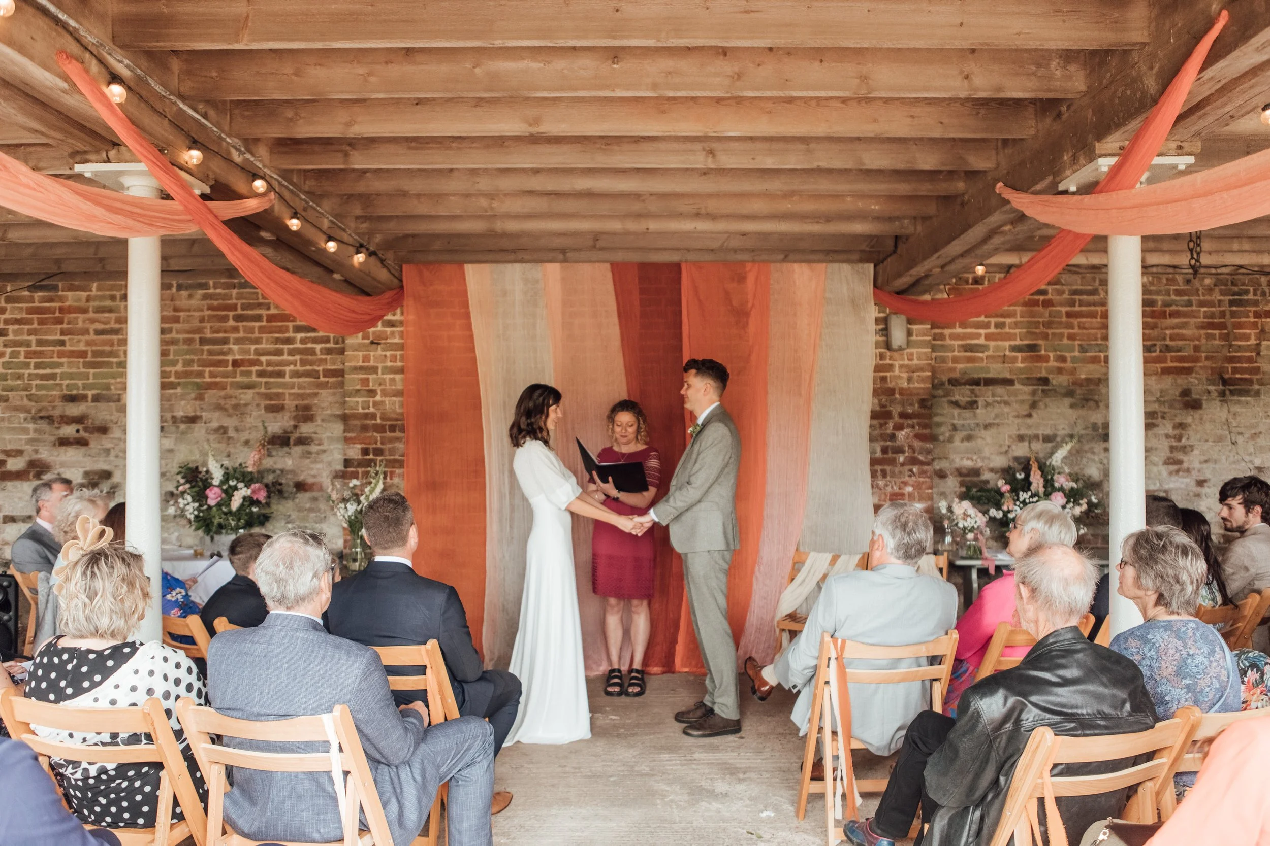 A romantic outdoor wedding ceremony set beneath rustic wooden beams at a countryside barn venue. The couple stands hand-in-hand in front of a beautiful fabric backdrop featuring soft terracotta, blush, and neutral drapes, creating a warm, earthy foca