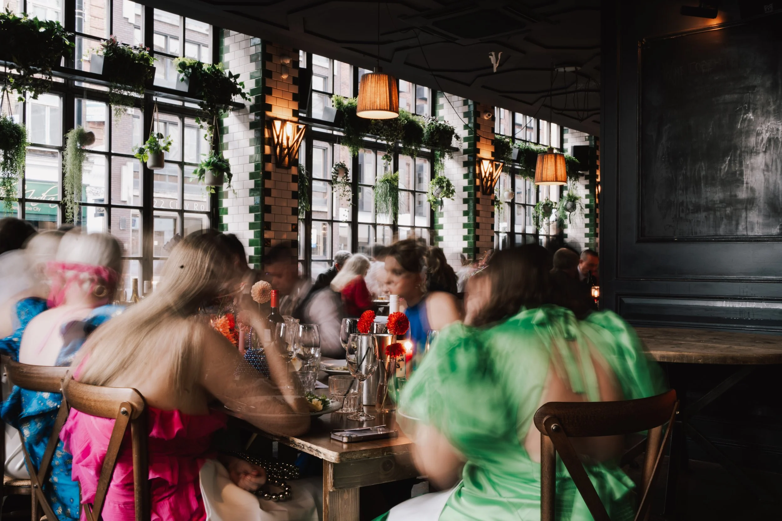 A lively wedding reception unfolds inside a stylish, plant-filled restaurant, where guests gather around wooden tables beneath warm hanging lamps and tall industrial windows. The soft motion blur captures the energy of conversation, laughter, and cli