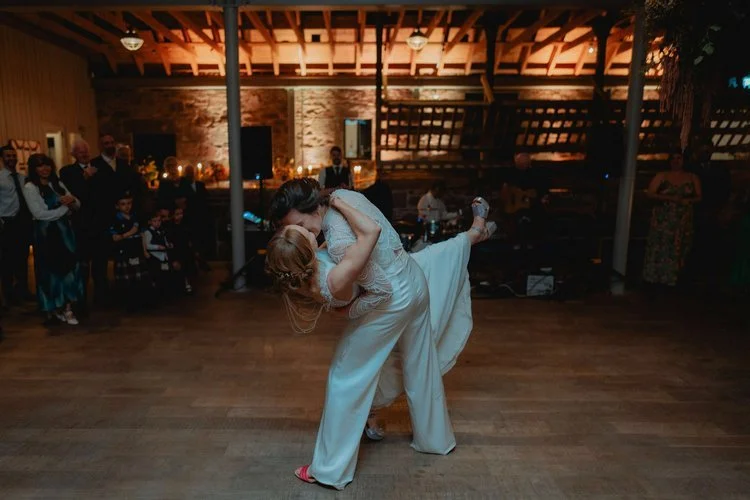 A breathtaking first-dance dip captured with cinematic flair by F.D Young Photography. One bride leans back in a dramatic swoop while the other holds her close, both completely lost in the music and the moment. The warm, rustic barn setting glows beh